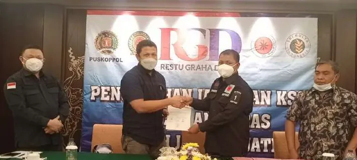 Dua pria bermasker berjabat tangan sambil memegang dokumen di depan spanduk bertuliskan RGD, disaksikan dua pria lain.