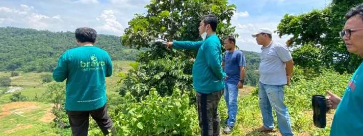 Lima pria berada di lereng bukit dengan latar hamparan lahan dan perbukitan; beberapa berkaus hijau toska, salah satunya bertuliskan bravo di punggung, dan seorang menunjuk ke arah lahan.