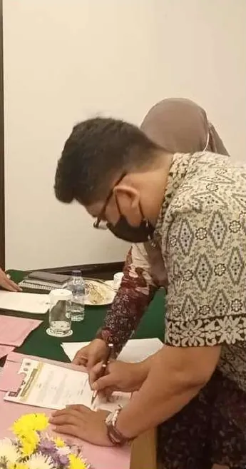 Seorang pria berkemeja batik dan bermasker menandatangani dokumen di meja berhias bunga; seorang perempuan berkerudung berada di belakangnya.