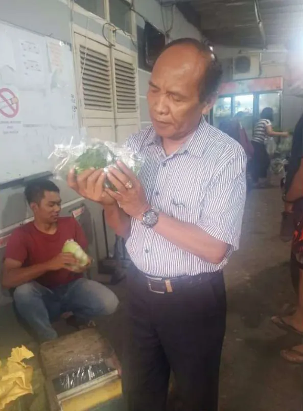 Seorang pria berkemeja garis memeriksa sayuran daun dalam kemasan plastik, sementara seorang pemuda berkaus merah duduk di lantai memegang sayuran.