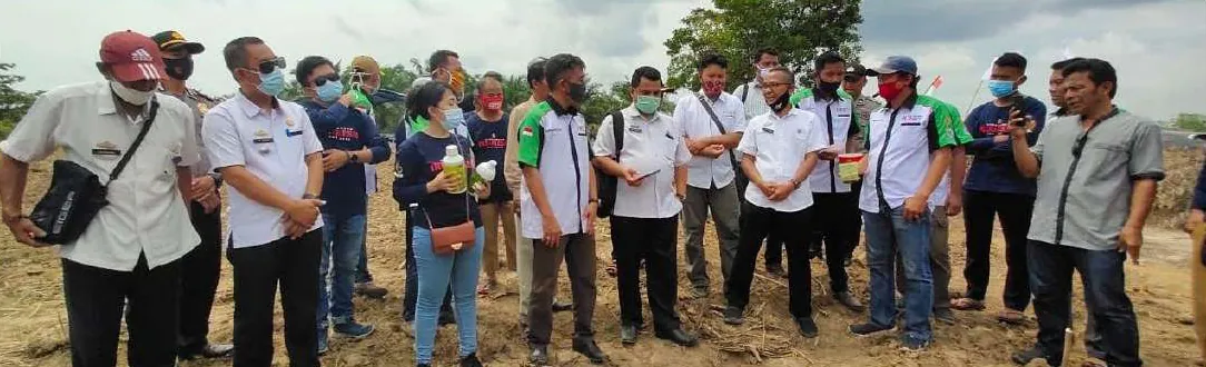 Rombongan orang, sebagian besar bermasker, berdiri di lahan terbuka bertanah kering; seorang perempuan memegang dua botol berlabel hijau.