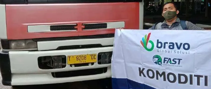 Seorang pria bermasker membentangkan spanduk putih bertuliskan "bravo Global Solusi", "FAST", dan "KOMODITI" di samping bagian depan truk merah-putih.