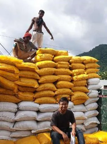 Tumpukan tinggi karung kuning dan putih di udara terbuka; dua pria berada di puncak tumpukan dan seorang pria duduk di depannya, dengan perbukitan di latar belakang.